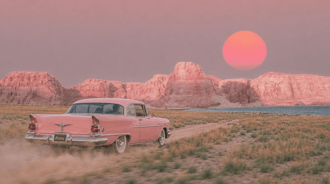 A pink vintage car driving through a desert at sunset